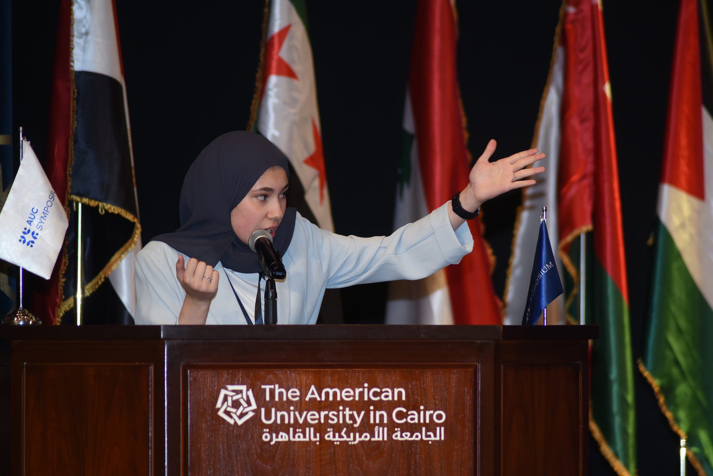 A debater speaking at the podium of The American University in Cairo with the flags of the region behind her